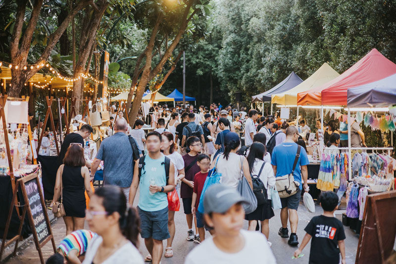 Crowded outdoor market with people browsing colorful stalls under trees. Strings of lights decorate the area, creating a lively atmosphere.