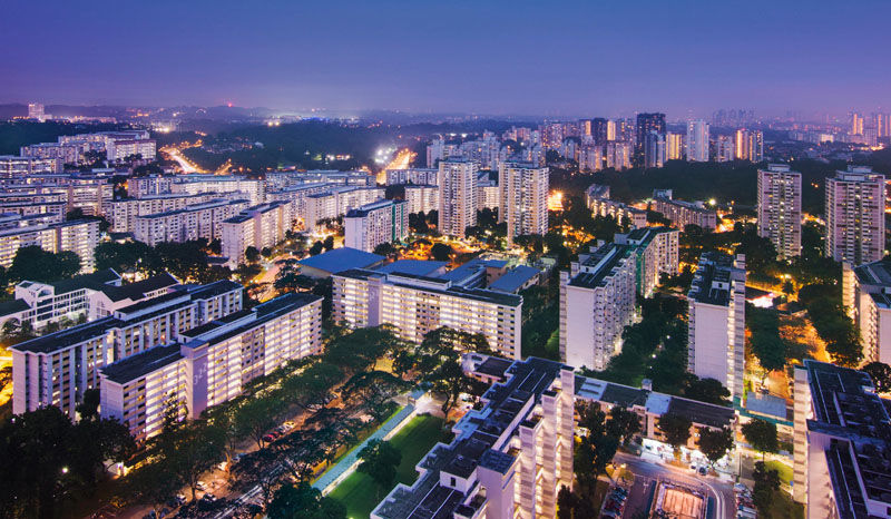 Cityscape at twilight with illuminated buildings, trees, and roads. Distant hills and a blue-purple sky create a tranquil urban atmosphere.