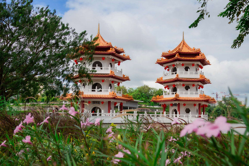 Two traditional pagodas with orange roofs stand amid lush greenery and pink flowers under a cloudy sky, creating a peaceful scene.