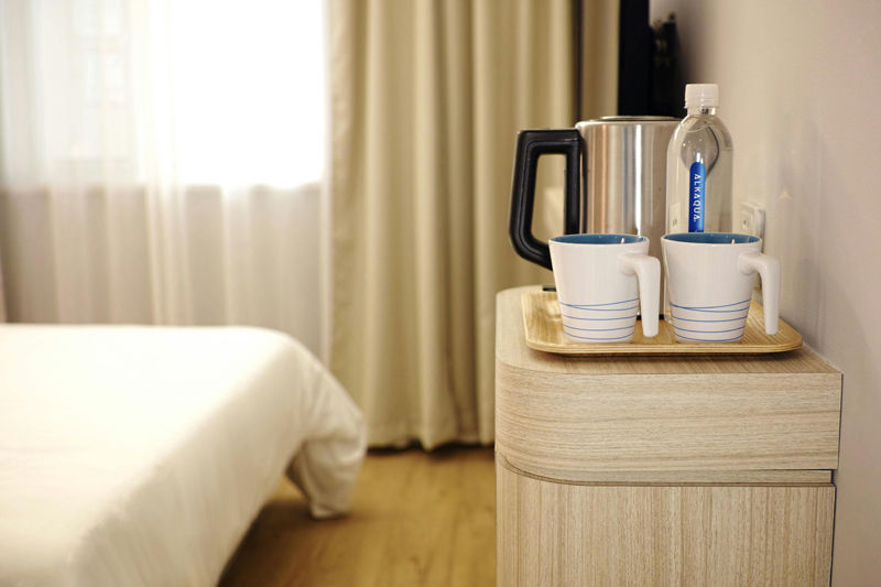 Tray with two striped mugs, kettle, and water bottle on a wood cabinet in a minimalist room with beige curtains and bed, creating a cozy vibe.