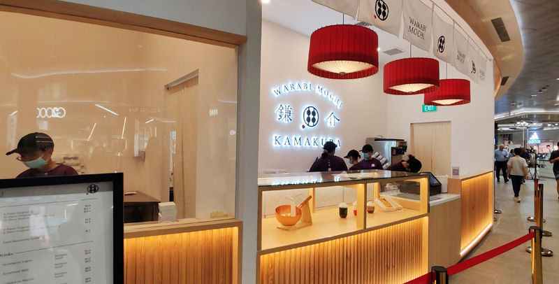 Employees work behind a counter at a Kamakura Warabi Mochi shop with glowing signage. Red lanterns hang above, and menus are visible.