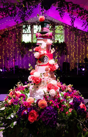 Alice in Wonderland themed wedding cake. 8 tiers tall. There is a top hat in green with a red sugar rose at the top of the cake. The 2nd and 3rd tiers are books. Next there is a white cake with a key hole and key. The 5th tier has a backwards clockface on it surrounded by gold. The 6th tier has cogs in bronze and gold. The 7th tier has playing cards featuring Alice, the hare and the Queen of Hearts. The bottom tier is covered in ruffles to represent Alice's skirt. It is surrounded by beautiful fresh flowers in pinks and purples.