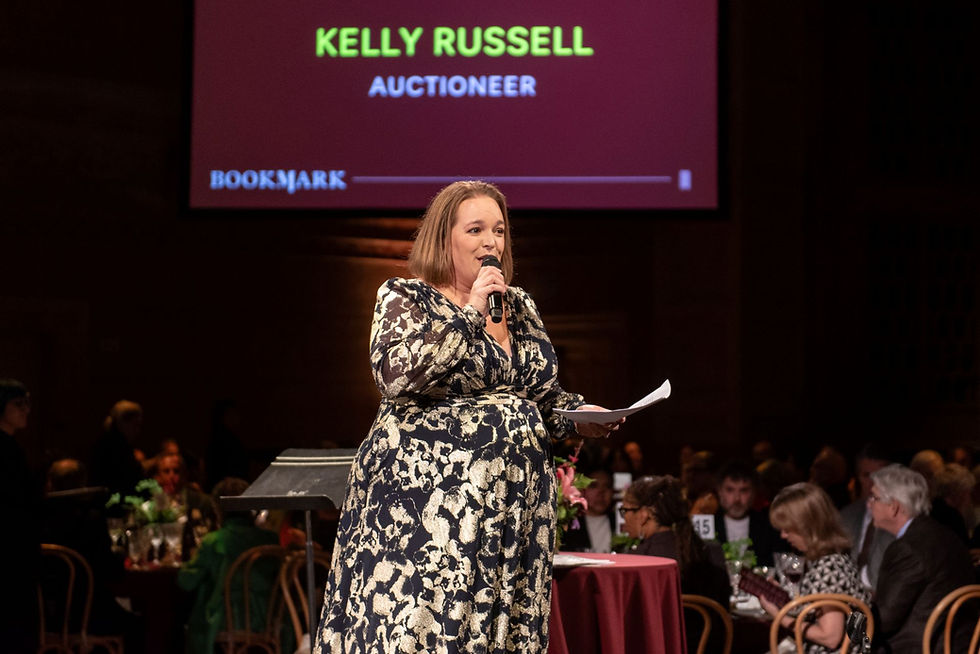 Benefit auctioneer Kelly Russell speaking on stage with a microphone in a dark room full of people with a banner featuring her name in the background.
