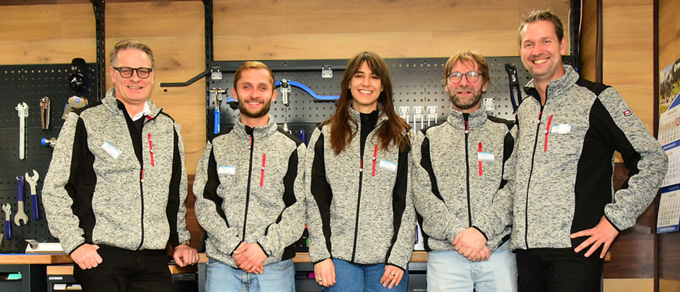 Das Team von der Fahrradwerkstatt RadFit, bestehend aus vier Männern und in der Mitte eine Frau stehen in Arbeitskleidung vor einer Werkbank.