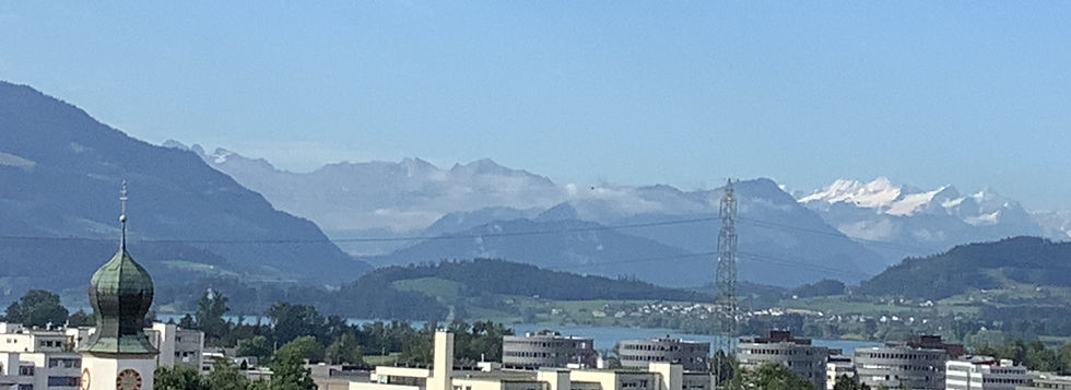Lake Zug waterfront with historic buildings, church tower, small boats, and Swiss mountains in the background.