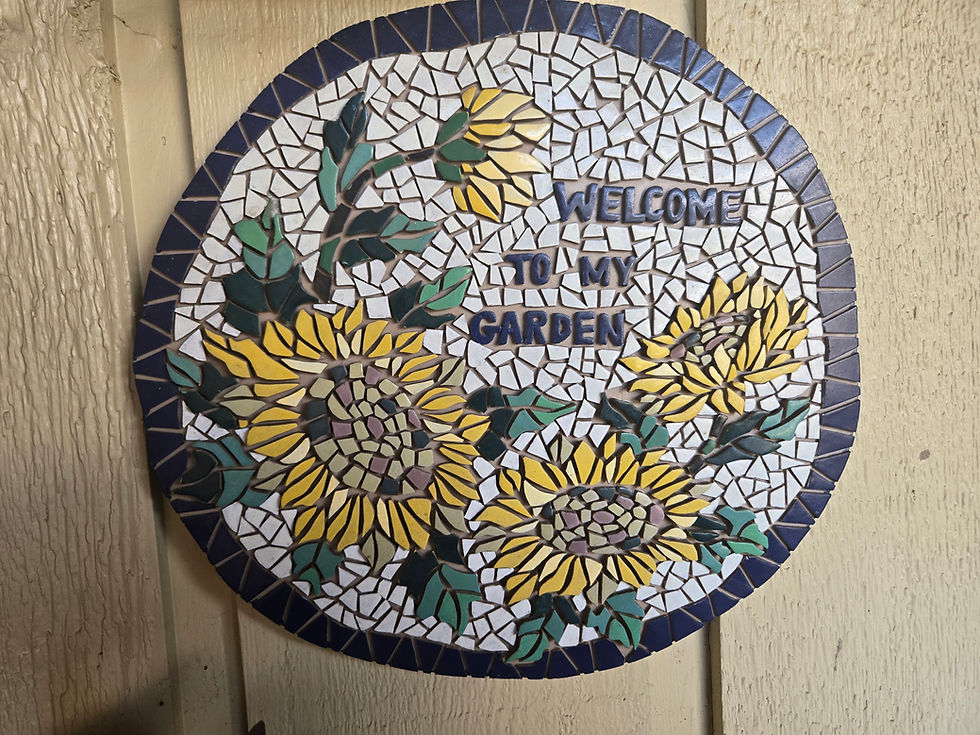Round sunflower mosaic sign on a beige wall reads WELCOME TO MY GARDEN, with yellow flowers and green leaves.