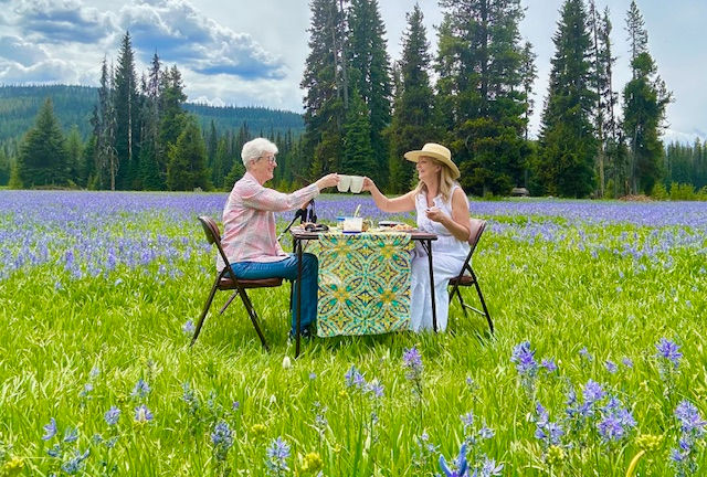 Two people clink cups at a table in a field of purple flowers. A colorful patterned cloth covers the table. Evergreen trees and mountains are in the background.