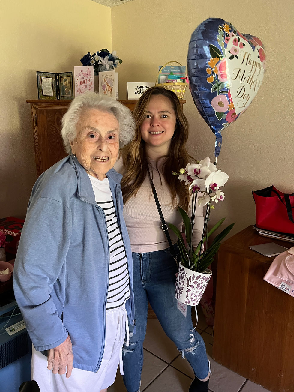 Elderly woman and young woman smiling in a home setting, holding a "Happy Mother's Day" balloon and flowers. Warm, cheerful atmosphere.