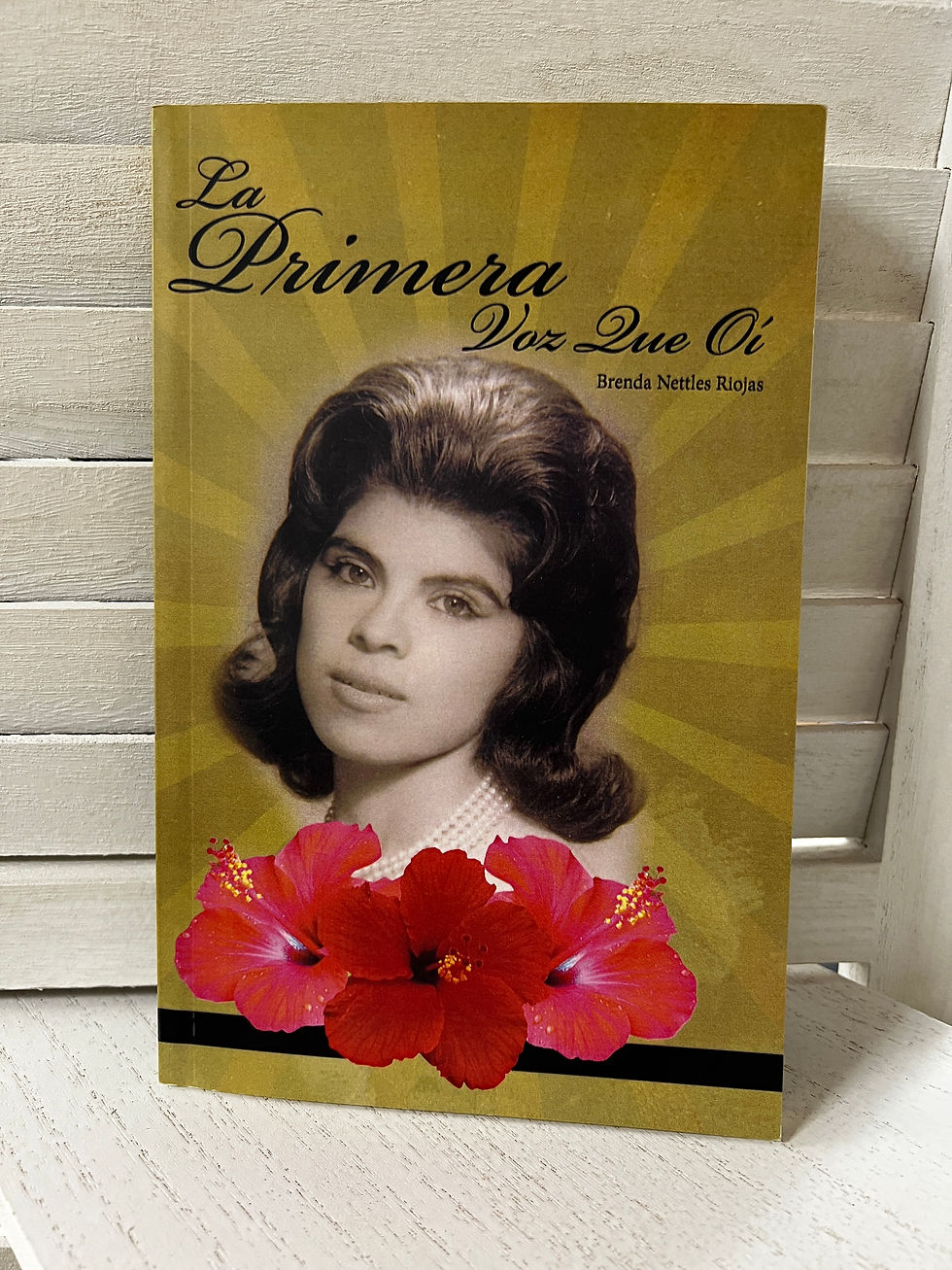 Book cover features a black-and-white portrait of a woman with a pearl necklace, three red hibiscus flowers, and text "La Primera Voz Que Oí".