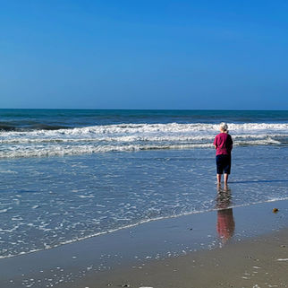 Elderly woman in pink shirt stands in shallow surf, watching waves on a sunny blue-sky beach.