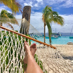 Relaxing in a hammock on a sandy beach, with palm trees, turquoise water, sailboats, and a bright blue sky in the background.