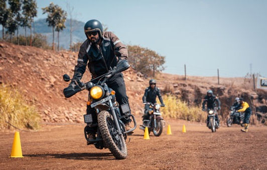 A person rides motorcycle, others behind them on dirt road with cones.