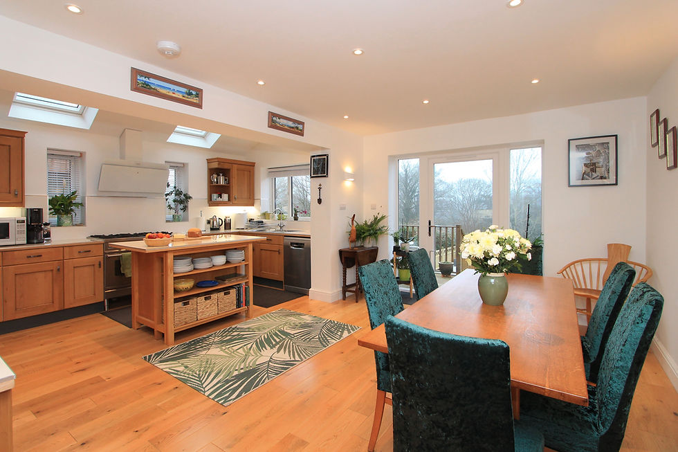 Bright open-plan kitchen and dining room with wood floors, teal chairs, island, flowers, skylights, and sliding doors to a deck.