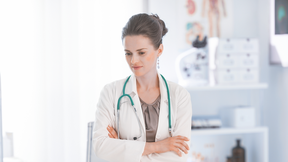 Female doctor with folded arms, wearing a white coat and stethoscope, appears thoughtful in a bright, medical office setting.
