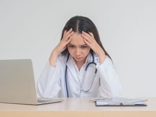 A stressed doctor in a white coat with a stethoscope, sitting in front of her laptop — a visual cue of burnout.