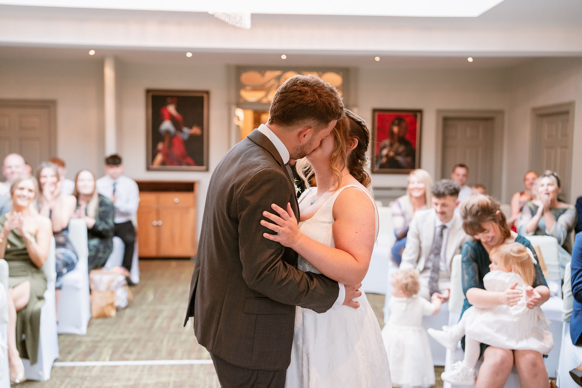 Bride and groom share an emotional kiss during their wedding ceremony, surrounded by smiling guests in an elegant wedding venue with classic artwork on the walls.