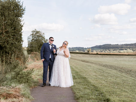 Bride and groom in sunglasses pose playfully on a country path. Bride in white dress, groom in blue suit. Sunny day with fields behind.