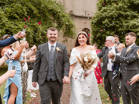 Bride and groom walk hand in hand, smiling, amid a celebratory confetti shower. Guests cheer in elegant attire, surrounded by greenery.