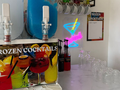 Slush machine labeled Frozen Cocktails on a counter with glasses. Neon cocktail sign in background. Bright, colorful decorations.