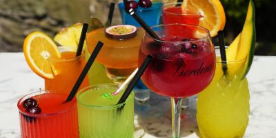 A vibrant assortment of frozen cocktails displayed on a marble surface, featuring a variety of colors including red, orange, green, yellow, and blue. Each drink is garnished with fresh fruits such as orange slices, cherries, passion fruit, and mango, and served in different styles of glasses with black straws. The background is blurred, showing an outdoor setting with natural lighting.