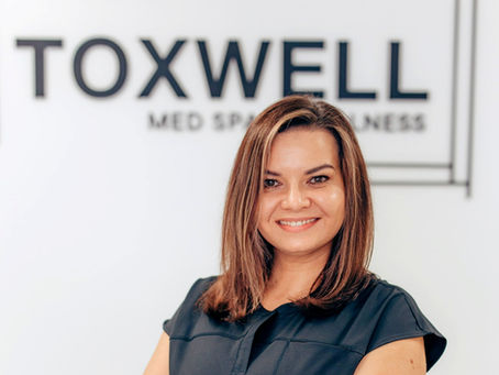 Smiling woman in black top stands in front of a sign reading "TOXWELL MED SPA & WELLNESS" on a white background.