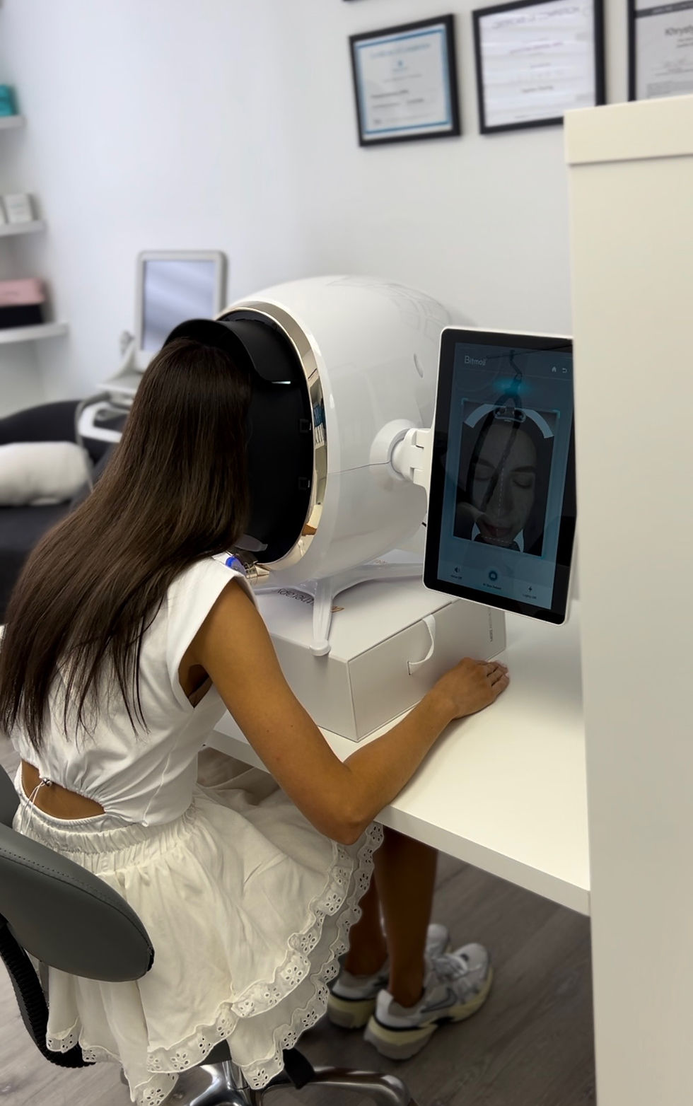 A woman in a white dress uses a facial scanner in an office. A tablet displays her image. The room has certificates on the wall.
