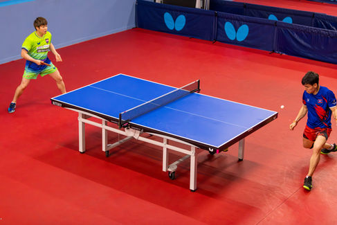 Table tennis players Hiromitsu Kasahara (left, in green) and Johan Hagberg (right, in blue) competing in a match at the ATTC Mega Money Open. Hagberg lunges to return the ball across a blue Butterfly table on a red court.