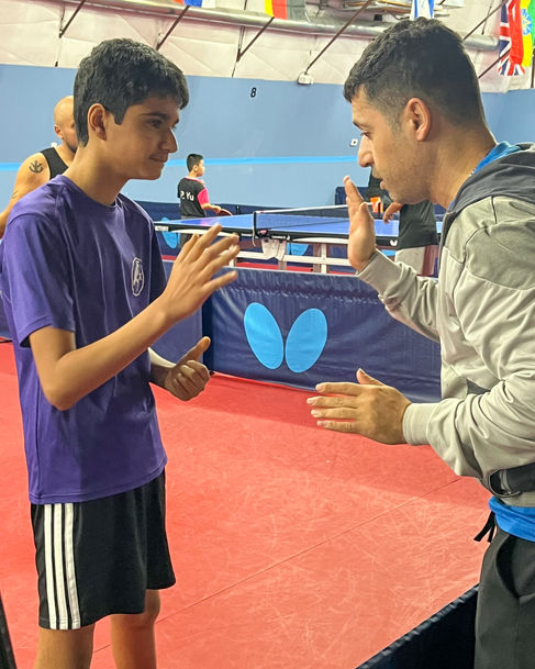 A young table tennis player in a purple shirt and black shorts gives a high-five to a coach, likely Coach Shahin Akhlaghpasand of the Austin Table Tennis Club. They are standing on red athletic flooring in a table tennis facility, with blue barriers and playing tables visible in the background.