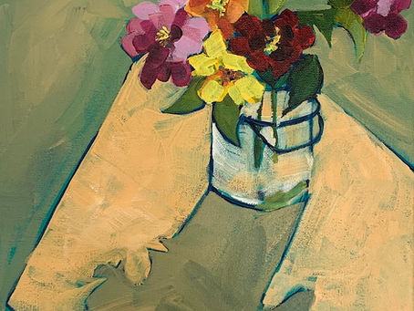 Vibrant flowers in a glass jar on a light wooden table. Green background, with shades of orange, pink, red, and yellow blooms.