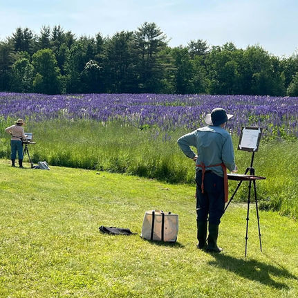 Artists painting in a grassy field with vibrant purple flowers. Sunlit scene with trees in the background, capturing a serene summer day.
