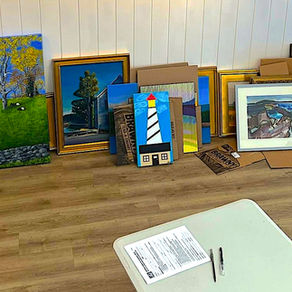 Framed paintings lean against a white wall; a table holds supplies. Bright landscapes and abstract art create a vibrant display.