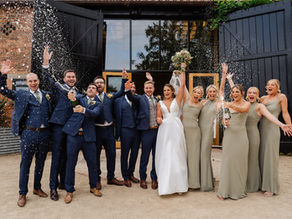 Bridal party joyfully sprays champagne, celebrating in front of a rustic venue. The bride is in white, surrounded by bridesmaids in sage dresses.