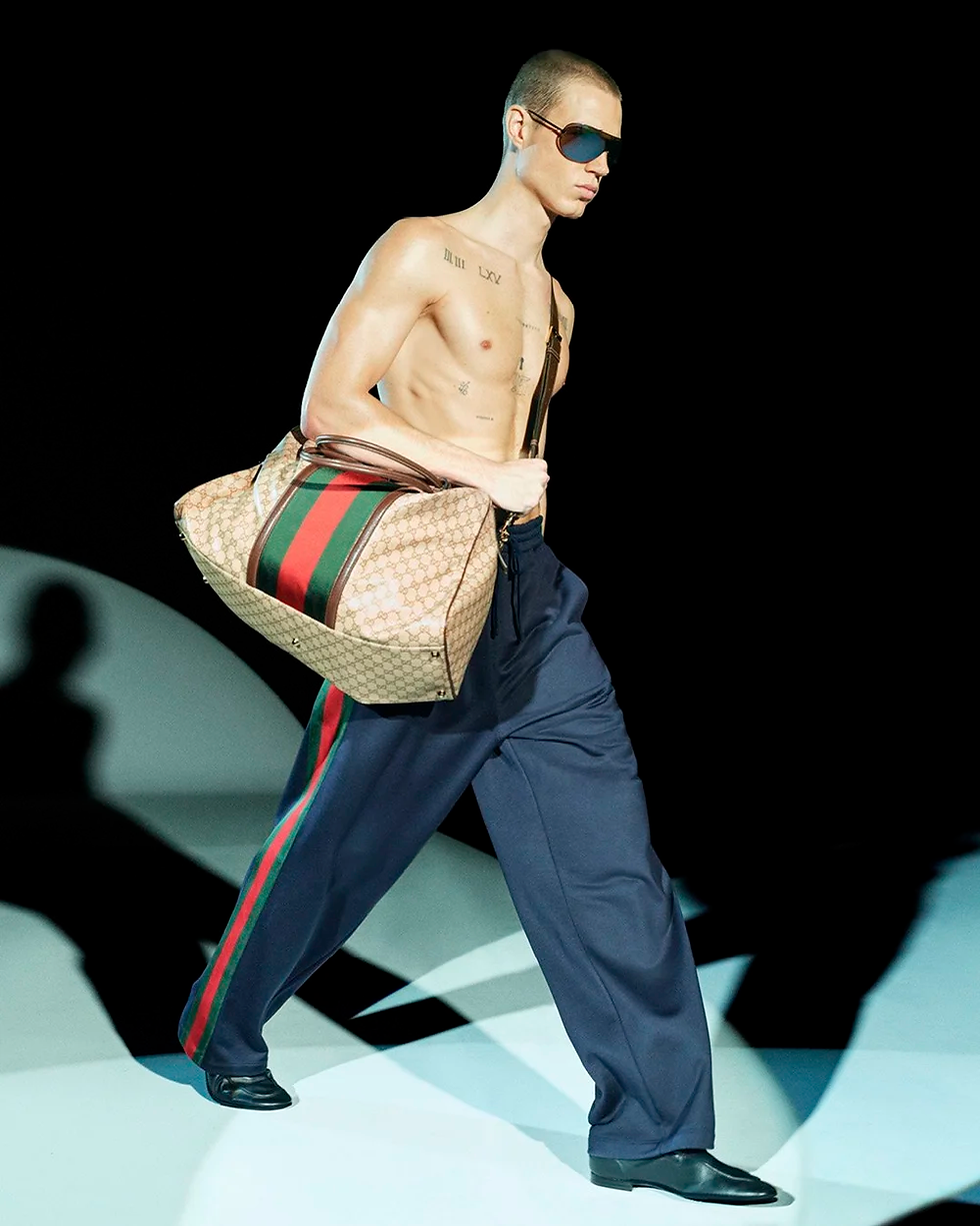 Shirtless model in sunglasses walking with an oversized beige Gucci monogram duffle bag and navy wide-leg trousers featuring red-and-green side stripes against a dark runway background.