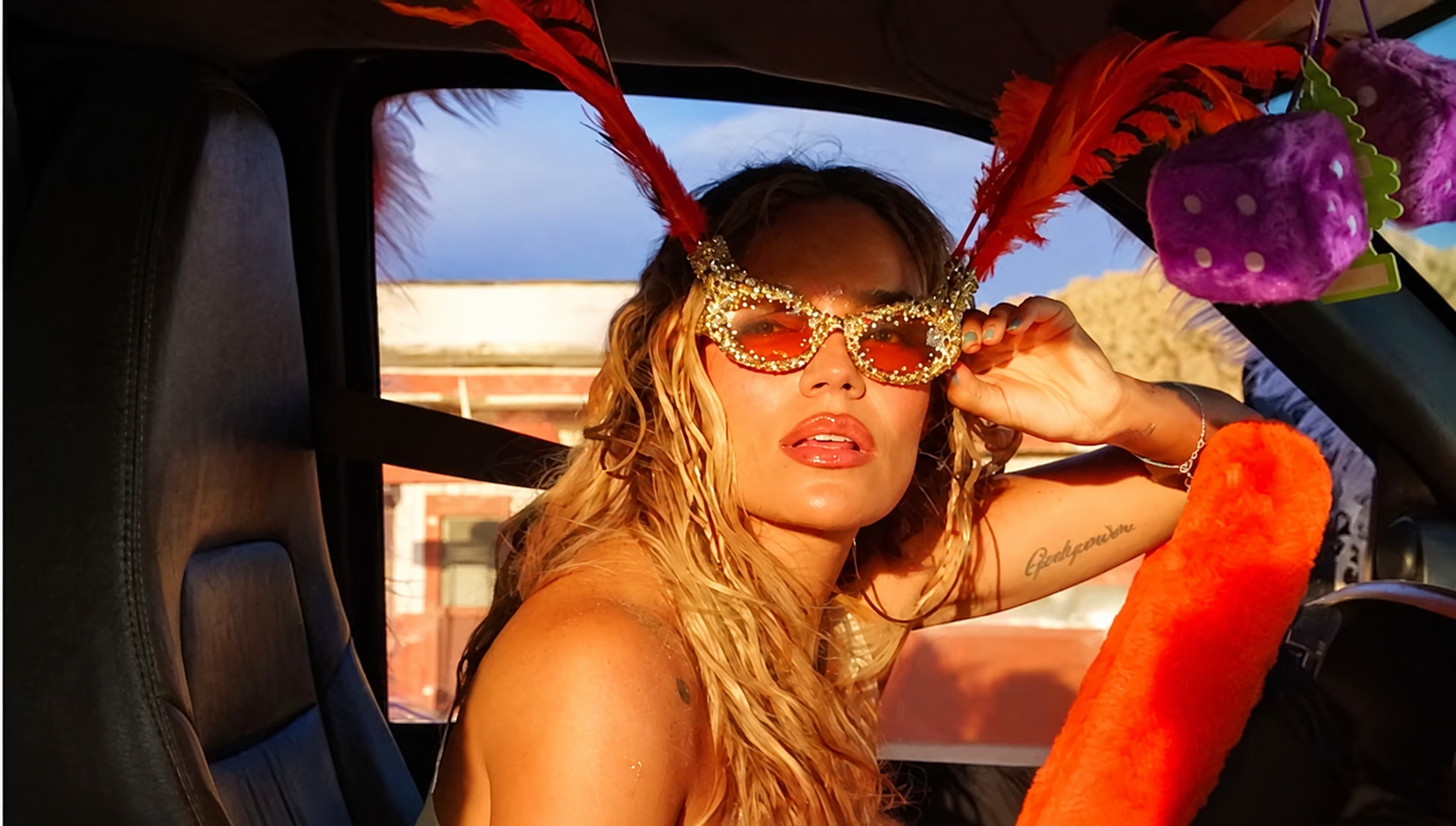 Karol G posing in a car wearing sparkling orange sunglasses with dramatic red feathers.