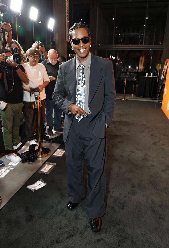 A$AP Rocky poses confidently on the red carpet in a loose-fitting navy suit with a polka dot tie and dark sunglasses, smiling as photographers capture his look at the Highest 2 Low screening.