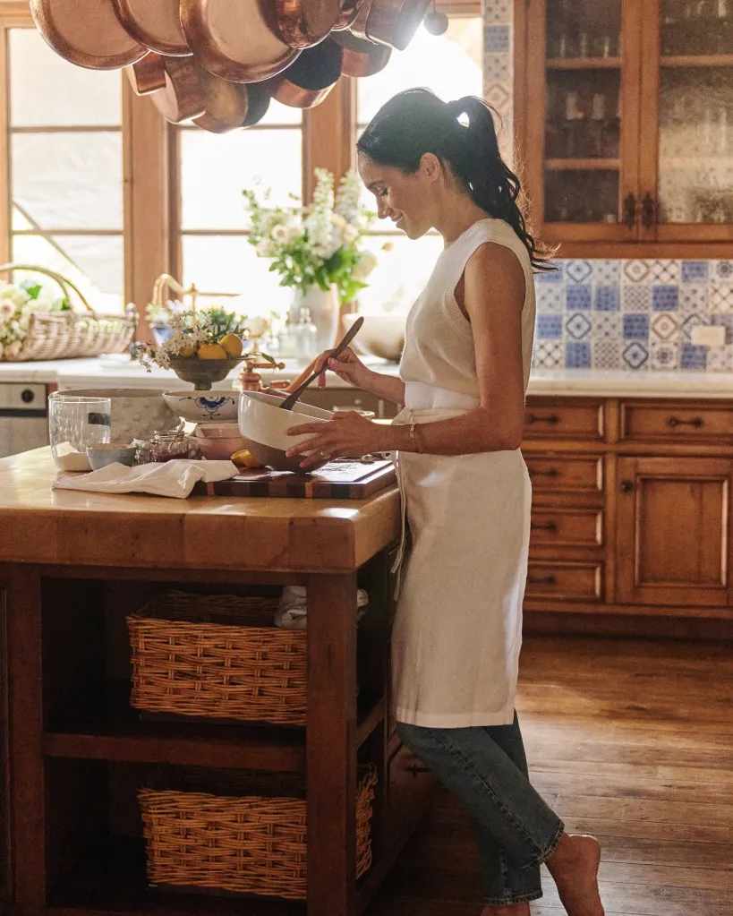 Meghan Markle cooking in a warm, rustic kitchen with wood cabinets, a hanging pot rack, and decorative tile backsplash.