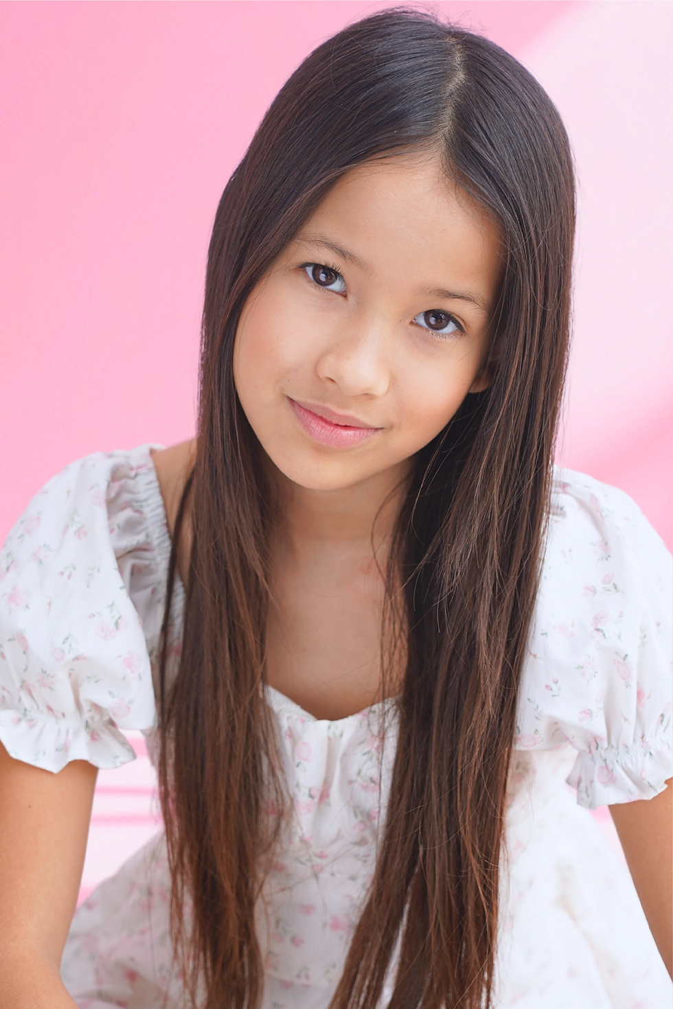 Portrait of Soleil River Nation in a white floral dress, leaning forward slightly and looking at the camera against a soft pink background.