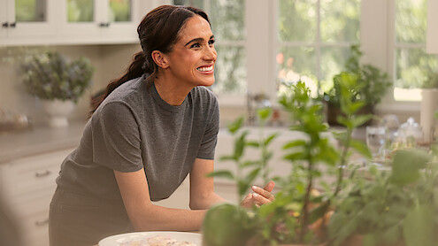 Meghan Markle smiling in a kitchen surrounded by greenery, during a scene from her Netflix show, highlighting her lifestyle and cooking content.