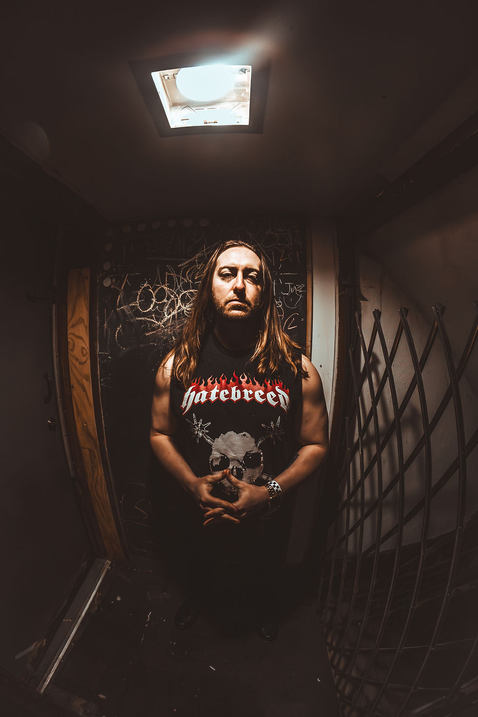 Kevin Powell standing in a dimly lit backstage-style room wearing a Hatebreed sleeveless shirt, with dramatic overhead lighting and dark walls behind him.