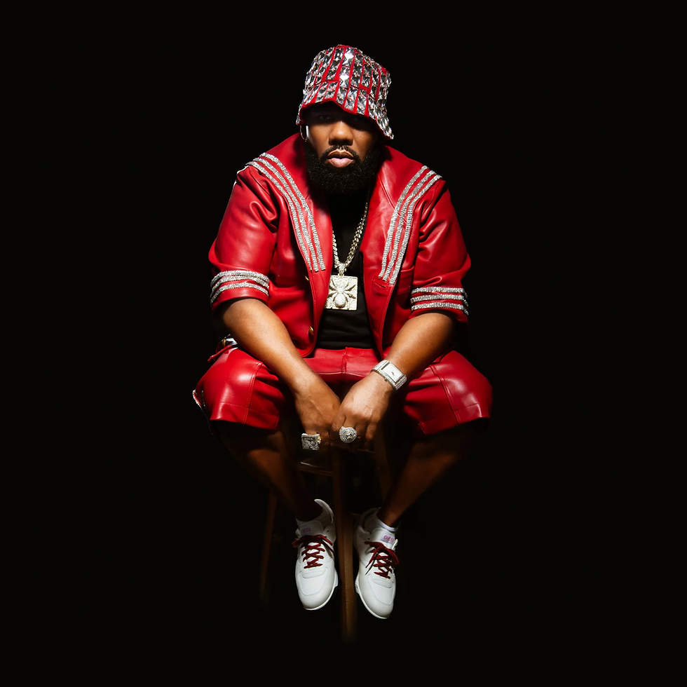 Raekwon wearing a red leather outfit and matching bucket hat, seated on a stool against a black background, styled with bold jewelry and white sneakers.