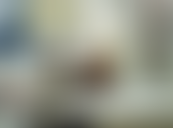 Scene from Messy Lola featuring Cynthia Kaye McWilliams seated at a modern office desk, leaning back in her chair with one leg extended onto the desk while holding a phone. Two table lamps frame the space, with a desktop computer and contemporary wall art visible in the background, conveying a relaxed yet slightly overwhelmed workplace moment.