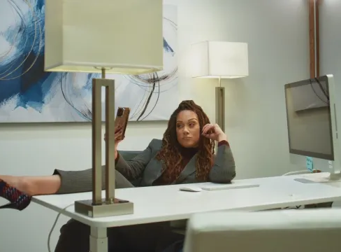Scene from Messy Lola featuring Cynthia Kaye McWilliams seated at a modern office desk, leaning back in her chair with one leg extended onto the desk while holding a phone. Two table lamps frame the space, with a desktop computer and contemporary wall art visible in the background, conveying a relaxed yet slightly overwhelmed workplace moment.