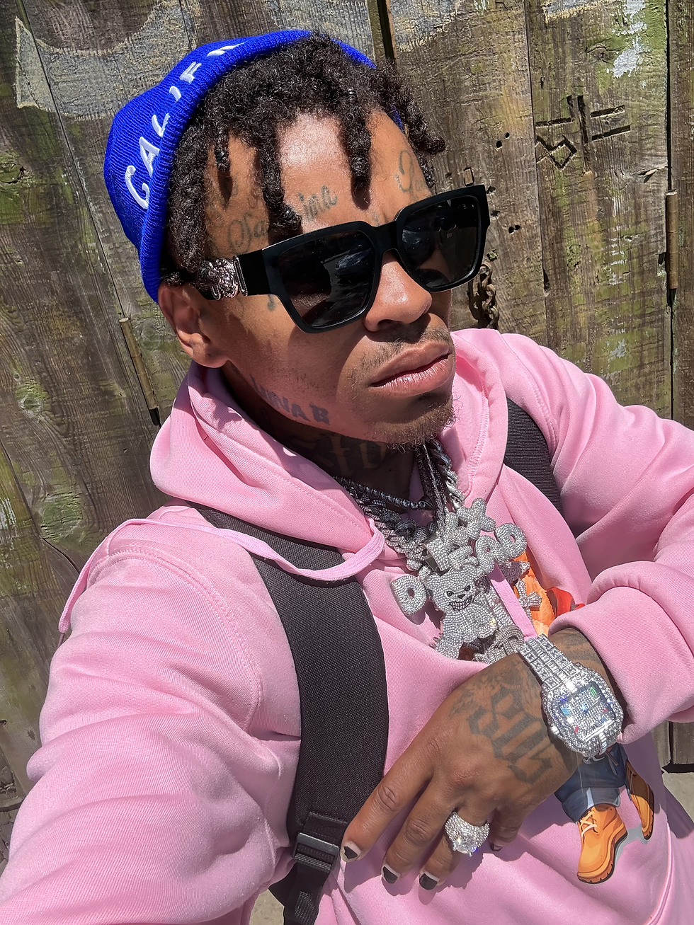 Young Drew wearing a pink hoodie, blue beanie, black sunglasses, diamond jewelry, and a watch while posing outdoors in front of a wooden fence.
