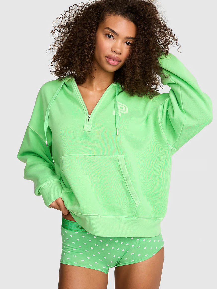 Model wearing a bright green PINK quarter-zip oversized hoodie with matching green heart-print boyshort underwear against a light gray background.