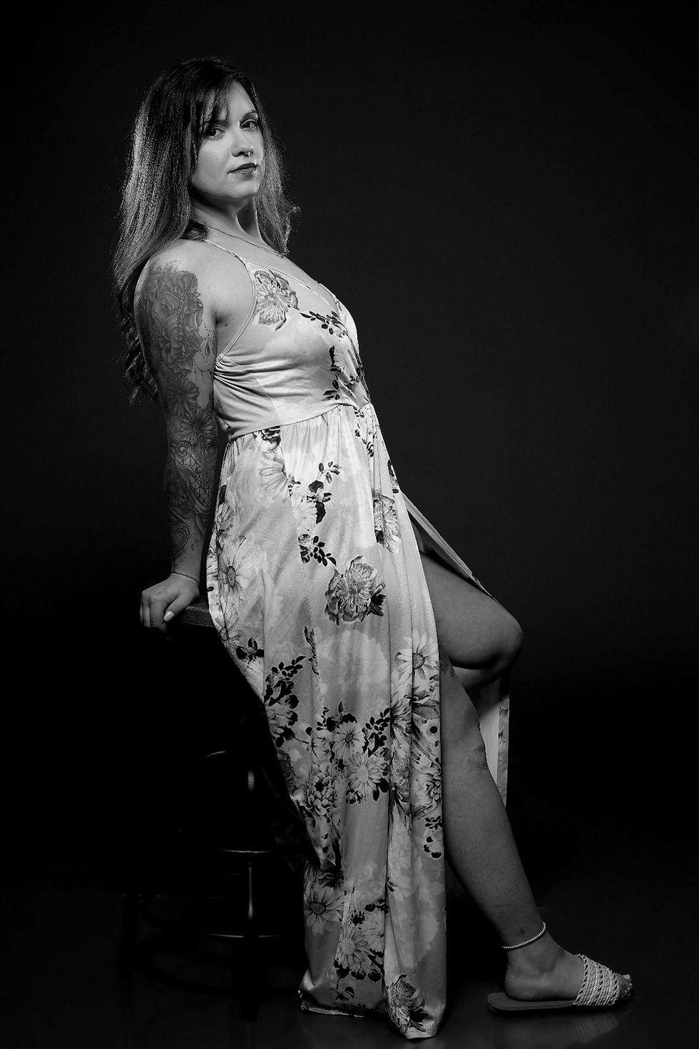Black-and-white full-body portrait of model Jaci Monroe posing beside a stool in a flowing floral dress, with her arm tattoos visible against a dark studio background.