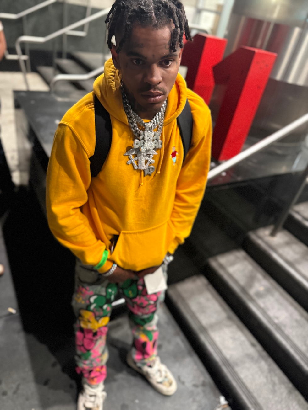 Young Drew wearing a yellow hoodie, colorful pants, white sneakers, a backpack, and layered jewelry while standing beside an indoor staircase.
