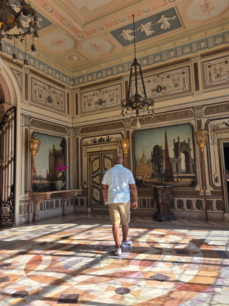 Dennis Nichols walks through an ornate historic interior with decorative ceilings, painted wall panels, chandeliers, and patterned marble floors. Vizcaya Museum & Gardens Miami.