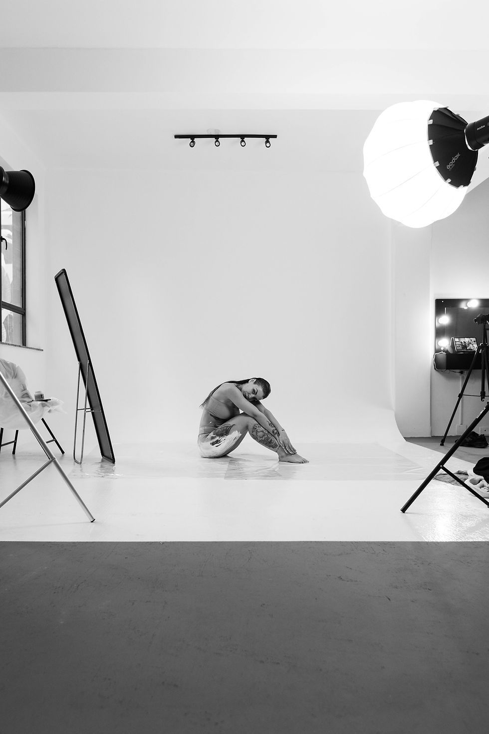 Black-and-white behind-the-scenes studio photo of a model seated on a white cyclorama backdrop during a photoshoot, surrounded by large softbox lights, mirrors, camera gear, and studio equipment.