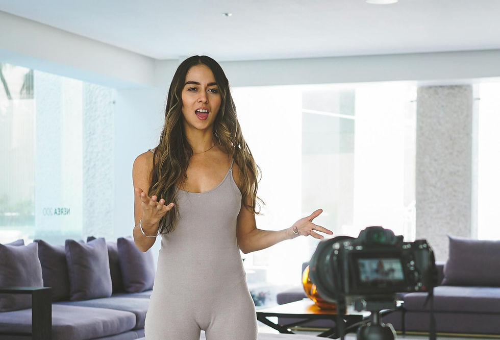 Woman in grey jumpsuit speaks energetically to a camera in a modern living room with purple couches and a yoga mat on the floor.