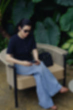 Woman wearing sunglasses, a black short-sleeve top, wide-leg jeans, and sandals sits on a woven outdoor chair surrounded by lush tropical plants, with a black Hermès-style handbag beside her.